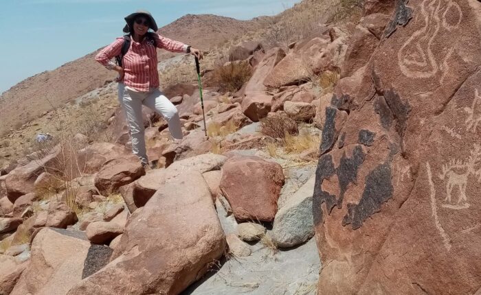 A woman leans on her walking pole on a hillside in desert terrain. There are petroglyphs on the rock beside her. Seen during a tour with Responsible Travel Peru from Arequipa