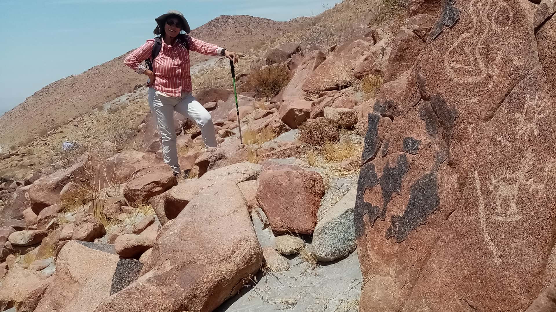 11A woman leans on her walking pole on a hillside in desert terrain. There are petroglyphs on the rock beside her. Seen during a tour with Responsible Travel Peru from Arequipa