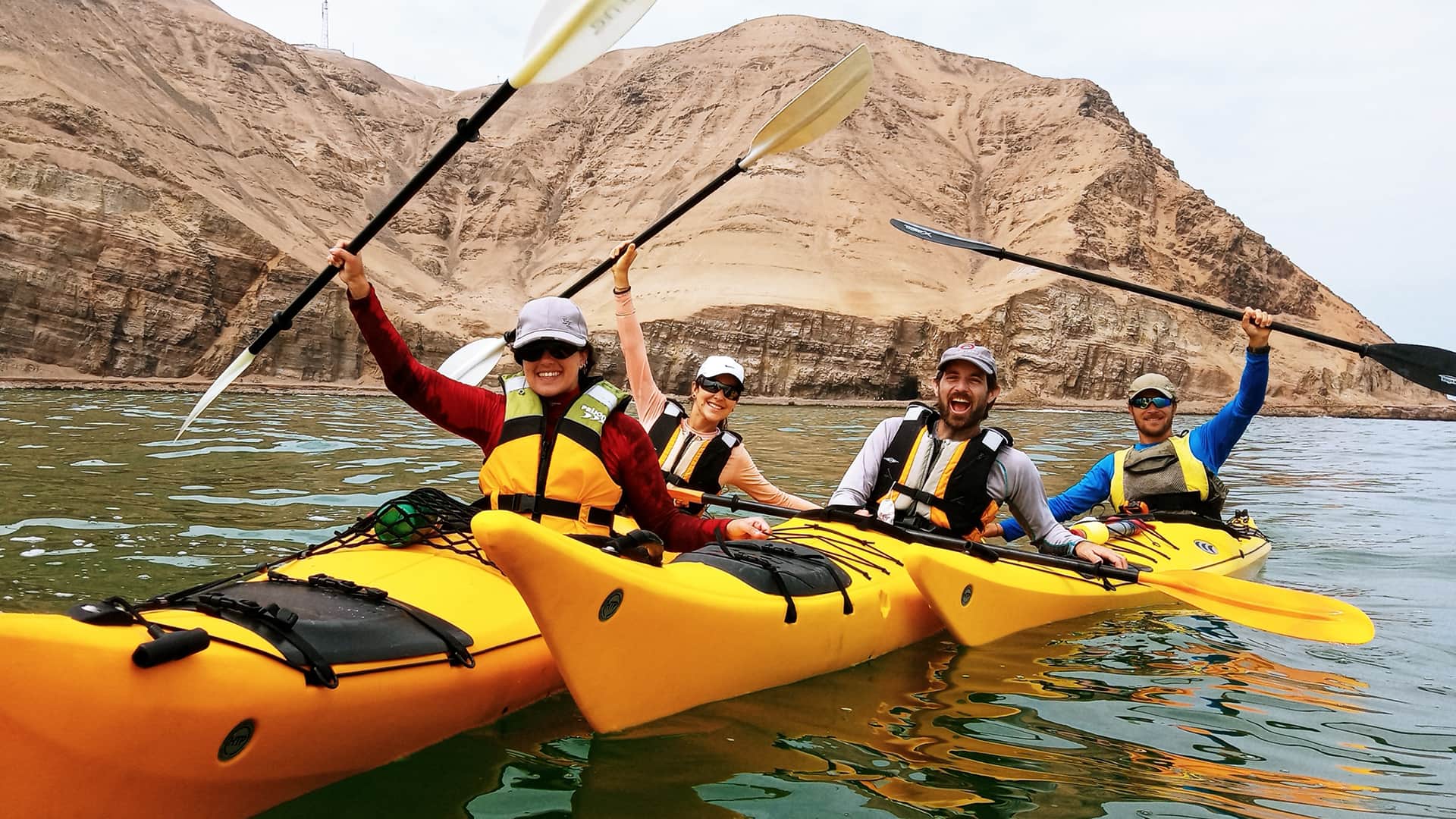 11four kayakers in three kayaks saluting with paddles up in Paracas bay where the sea meets the the desert | Responsible Travel Peru